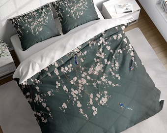 May include: A bedding set featuring a dark teal duvet cover and two pillowcases. The design includes a floral pattern with white blossoms and small blue birds. The bedding is on a white bed with a white sheet.