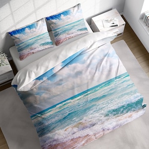 May include: A white duvet cover with a blue and white ocean wave design. The duvet cover features a close-up of the ocean with white foamy waves crashing on the shore. The sky is a light blue with white clouds.