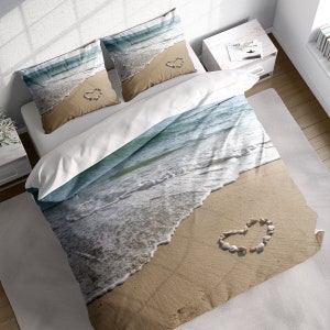 Peut inclure: Une housse de couette blanche avec un motif de plage imprimé. La scène représente des vagues bleues de l'océan qui se brisent sur une plage de sable avec un cœur en forme de coquillages.