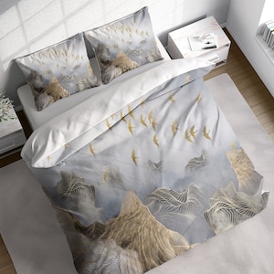 May include: A white and grey duvet cover with a gold mountain and bird design. The duvet cover is on a bed with two matching pillowcases.
