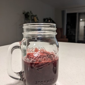 Puede incluir: Un vaso de vidrio transparente tipo tarro Mason lleno de un líquido morado oscuro, posiblemente jugo o un batido. El vaso tiene un asa y la palabra "STORAGE" está grabada en el lateral. El líquido está cubierto con una espuma rojiza.