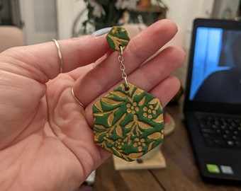 May include: A pair of green and gold hexagonal earrings with a floral pattern. The earrings are made of polymer clay and have a silver chain.