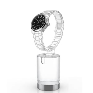 Peut inclure: Un présentoir de montre en acrylique transparent avec une base argentée, présentant une montre-bracelet avec un cadran noir et un bracelet argenté. La montre est suspendue verticalement, mettant en valeur son design. Le support est cylindrique.