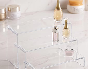 May include: A clear acrylic three-tiered display stand with a stepped design. The stand features three rectangular shelves of varying heights, constructed from transparent material, providing a tiered surface for organizing small items.