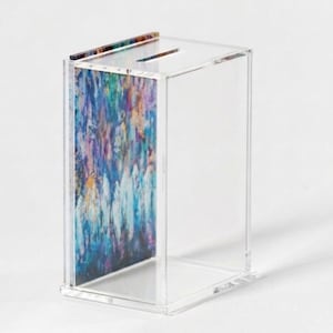 May include: Clear acrylic box with a slot on top for inserting items. The front panel features a colorful abstract design with shades of blue, purple, and white. The box is rectangular and appears to be a decorative storage item.