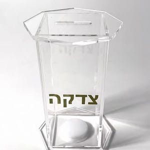 Könnte beinhalten: Transparente Spendenbox aus Acryl mit sechseckiger Basis und Oberseite. Die Box hat einen Schlitz für Spenden und das Wort "Tzedakah" in goldfarbenen Buchstaben. Ein weißes, rundes Objekt ist im Inneren sichtbar.