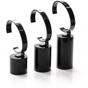 May include: Three glossy black watch display stands of varying heights, each featuring a cylindrical base and a curved C-shaped arm designed to hold a timepiece. The stands are arranged in a row, showcasing a smooth, reflective finish throughout.