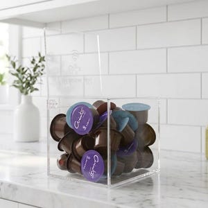 Op de afbeelding: Een heldere acryl koffiepadhouder gevuld met diverse gekleurde koffiepads. De houder is rechthoekig met een scharnierend deksel en staat op een wit aanrechtblad. De achtergrond omvat een witte tegelmuur, een witte vaas met groen en een groene fles.
