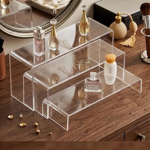 Op de afbeelding: Transparante acryl display risers in een trapsgewijze opstelling. De risers bevatten parfumflesjes, sieraden en een huidverzorgingsproduct. De opstelling staat op een houten oppervlak, met een spiegel en andere schoonheidsproducten op de achtergrond.