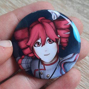 May include: A round button pin featuring a colorful anime-style illustration of a character with red hair and eyes. The character is wearing a gray and red outfit against a dark blue background. The pin is held in a hand.