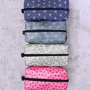 Könnte beinhalten: Ein Stapel rechteckiger Reißverschlusstaschen in verschiedenen Mustern. Die oberste Tasche ist marineblau mit einem weißen geometrischen Design. Darunter befindet sich eine Tasche mit einem grau-weißen Blättermuster, gefolgt von einer Tasche mit einem grün-weißen Blumendesign und einer rosa Tasche mit roten Erdbeeren.