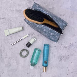 Pode incluir: Uma bolsa de cosméticos azul com um padrão de folhas brancas, aberta para revelar uma escova de cabelo de madeira. Ao redor da bolsa estão produtos de beleza, incluindo um tubo de creme, rímel, esmalte de unhas, um elástico de cabelo e duas garrafas de líquido.