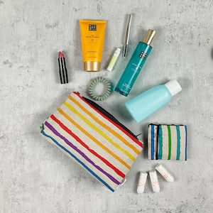 Könnte beinhalten: Eine gestreifte Stofftasche mit Reißverschluss, eine Flasche mit blauer Flüssigkeit, eine Flasche mit grüner Flüssigkeit, eine Tube gelber Lotion, ein Lippenstift, ein Haargummi, eine kleine Tube weißer Lotion, ein schwarzer Eyeliner und drei weiße Tampons.