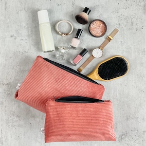 Pode incluir: Uma variedade de produtos de beleza e acessórios. Duas bolsas de cosméticos de veludo cotelê coral estão abertas. Outros itens incluem esmalte de unhas, um relógio, uma escova de cabelo, um pincel de maquiagem, uma pulseira e perfume.