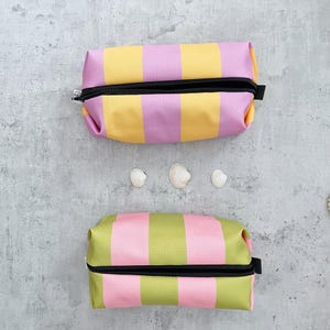 Könnte beinhalten: Zwei gestreifte Kosmetiktaschen. Eine Tasche hat gelbe und rosa Streifen, die andere hat grüne und rosa Streifen. Beide Taschen haben einen schwarzen Reißverschluss und einen Zipper. Die Taschen sind zylindrisch geformt.