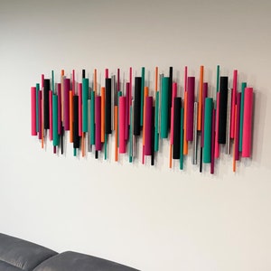 May include: A wall-mounted art installation consisting of numerous vertical cylindrical rods of varying lengths and diameters. The rods are painted in a vibrant color palette of magenta, teal, orange, black, and silver, arranged in a dense horizontal cluster.