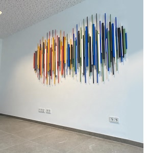 Könnte beinhalten: Abstrakte Wandkunst mit vertikalen Stäben in verschiedenen Farben, darunter Rot, Gelb, Blau und Grün. Die Stäbe sind in einer wolkenartigen Form an einer weißen Wand angeordnet und schaffen eine moderne und farbenfrohe Darstellung. Das Kunstwerk verleiht dem Innenraum eine lebendige Note.