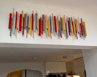 Op de afbeelding: Een wandkunstinstallatie bestaande uit talloze verticale cilindrische staven van verschillende lengtes en diameters. De staven zijn gerangschikt in een dicht, lineair patroon en hebben een kleurenpalet van rood, geel, grijs en koper.