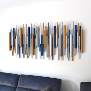 May include: Abstract wall art featuring vertical wooden rods in shades of navy blue, silver, and gold. The rods are mounted on a white wall above a navy blue sofa. This modern art piece adds a touch of color and texture.