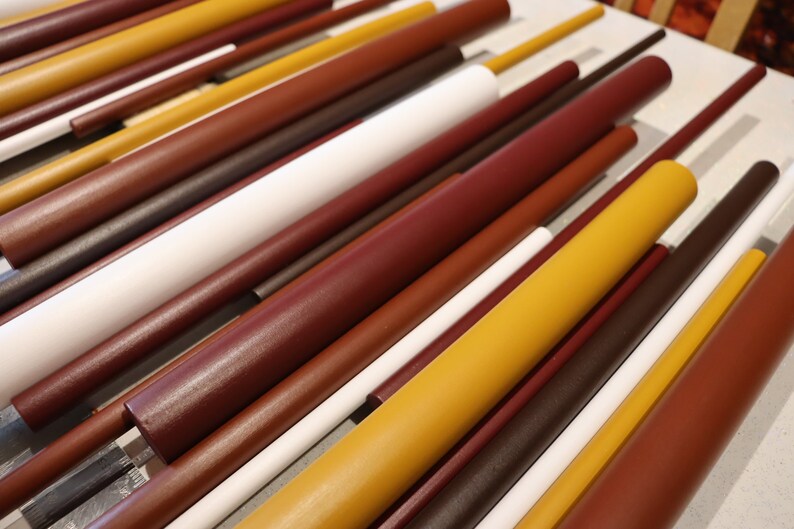 May include: A collection of cylindrical rods arranged in parallel, featuring a color palette of mustard yellow, deep burgundy, dark brown, and white. The rods vary in diameter and length, creating a layered, linear composition with smooth surfaces.