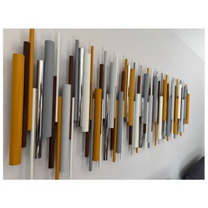 May include: A wall-mounted sculpture composed of numerous vertical cylindrical rods of varying lengths and diameters. The rods feature a color palette of mustard yellow, matte gray, white, dark brown, and reflective metallic silver finishes.