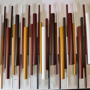 May include: A wall art piece composed of numerous vertical cylindrical rods of varying lengths and diameters. The rods are painted in a palette of white, mustard yellow, dark brown, and reddish-brown, arranged in a dense, staggered pattern.