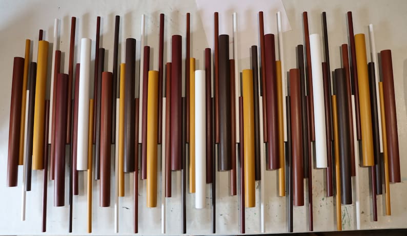 May include: A wall art piece composed of numerous vertical cylindrical rods of varying lengths and diameters. The rods are painted in a palette of white, mustard yellow, dark brown, and reddish-brown, arranged in a dense, staggered pattern.