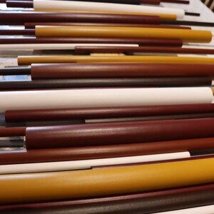 May include: A collection of cylindrical rods arranged horizontally in a parallel pattern. The rods vary in length and thickness, featuring a color palette of matte white, deep brown, dark maroon, and mustard yellow with smooth, solid finishes.