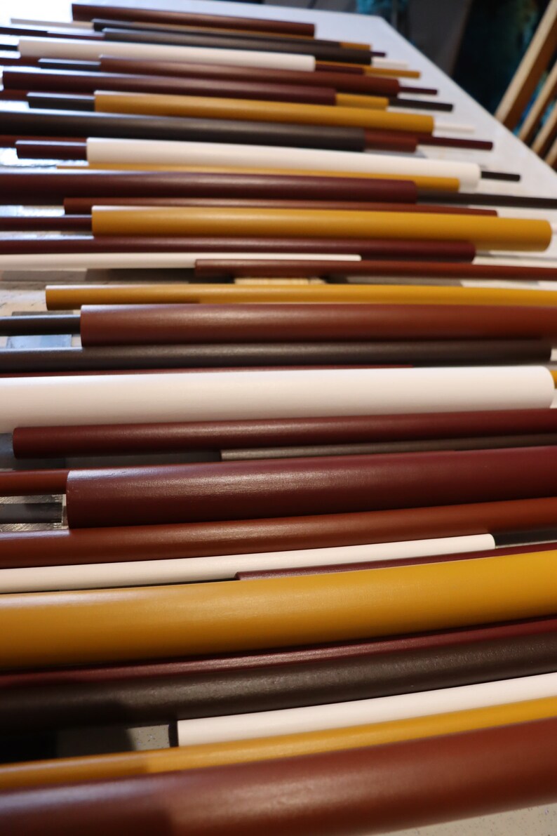 May include: A collection of cylindrical rods arranged horizontally in a parallel pattern. The rods vary in length and thickness, featuring a color palette of matte white, deep brown, dark maroon, and mustard yellow with smooth, solid finishes.