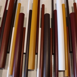 May include: A wall art piece composed of numerous vertical cylindrical rods of varying lengths and diameters. The rods are arranged in a parallel pattern and feature a color palette of mustard yellow, deep burgundy, dark brown, and crisp white.