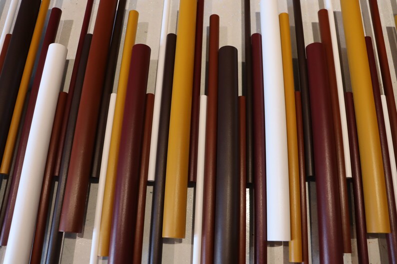 May include: A wall art piece composed of numerous vertical cylindrical rods of varying lengths and diameters. The rods are arranged in a parallel pattern and feature a color palette of mustard yellow, deep burgundy, dark brown, and crisp white.