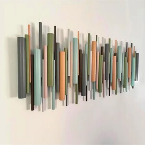 May include: A wall-mounted art installation composed of numerous vertical cylindrical rods of varying lengths and diameters. The rods are painted in a palette of muted tones including sage green, terracotta, charcoal grey, soft pink, and light blue.