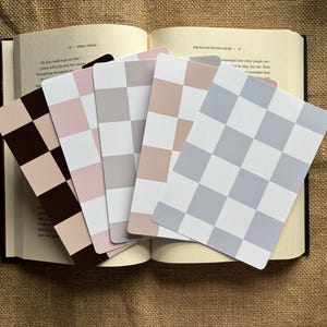 May include: An open book with several checkerboard pattern bookmarks in shades of brown, pink, grey, and white. The bookmarks are fanned out over the pages, with the book resting on a textured, tan surface.