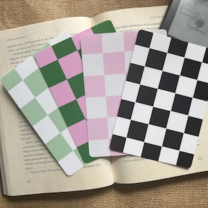 May include: An open book with several checkered pattern bookmarks in green, pink, and black and white. An e-reader is visible in the background. The bookmarks have a square pattern design.