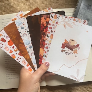 May include: A selection of autumn-themed bookmarks featuring designs of pumpkins, leaves, and ghosts. The bookmarks are displayed over an open book and a digital e-reader. The colour scheme includes shades of brown, orange, and white.