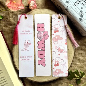May include: Three pink and white bookmark designs with pink tassels. The first bookmark has a pink cowboy boot and the text "Wear the Hat, Ride the Cowboy". The second bookmark has the word "Howdy" in pink letters. The third bookmark has a pink and white pattern of cowboy hats.
