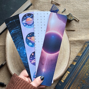 May include: Four different space-themed bookmarks. One bookmark features a galaxy design with a blue and purple colour scheme. Another bookmark features a planet with rings and a blue and orange colour scheme. A third bookmark features a galaxy with a black hole and a purple and pink colour scheme. The final bookmark features a galaxy with a black hole and a blue and pink colour scheme.