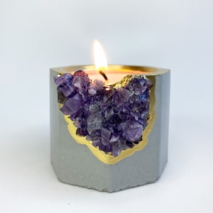 Pode incluir: Um pequeno porta-velas de concreto cinza com uma borda dourada e um aglomerado de cristais de ametista roxa dentro. A vela está acesa e a chama é visível.