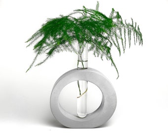 May include: A gray concrete vase with a circular opening, holding a glass test tube with green sprigs of asparagus fern.