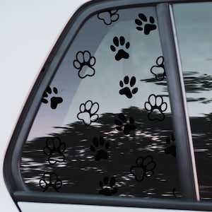 Puede incluir: Una ventana de coche con múltiples pegatinas negras con forma de huellas de pata. Las huellas varían en tamaño y están dispersas por el cristal. El fondo muestra una vista borrosa de árboles y cielo, sugiriendo un entorno exterior.