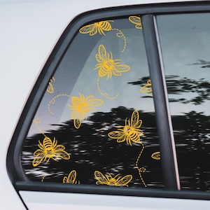 Może przedstawiać: Szyba samochodowa z wieloma żółtymi naklejkami z pszczołami. Pszczoły są w różnych fazach lotu, z przerywanymi liniami wskazującymi ich trajektorie. Tło przedstawia rozmyty widok drzew i nieba.