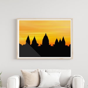May include: A framed print of a silhouette of a temple complex against a sunset sky. The sky is a gradient of orange and yellow. The temple is black and has a series of towers.