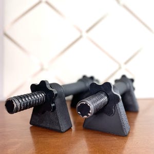 Könnte beinhalten: Schwarze Hantelgriffe mit sternförmigen Kragen. Die Griffe liegen auf einer Holzoberfläche. Der Hintergrund ist eine weiße Wand mit einem Rautenmuster.
