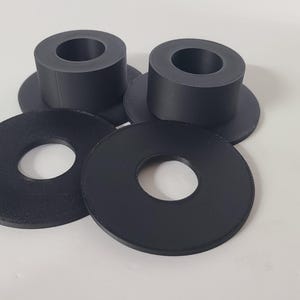 May include: Two matte black cylindrical spacers with wide circular flanges and two flat black circular washers with central holes. These four components are arranged in a cluster, featuring a smooth, uniform finish and a simple geometric design.