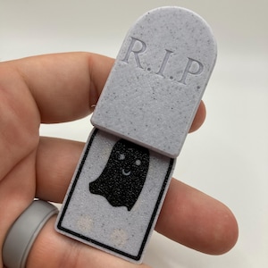 May include: A grey tombstone-shaped Halloween toy with the letters "R.I.P." on top. The bottom section reveals a black ghost with a smiling face. The toy is held in a hand.