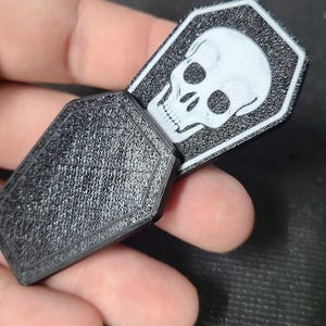 Pode incluir: Objeto preto em forma de caix&atilde;o com superf&iacute;cie texturizada. Um gr&aacute;fico de caveira branca &eacute; vis&iacute;vel em uma etiqueta retangular que desliza para fora do caix&atilde;o. A etiqueta tem uma borda preta.