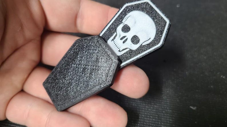 Pode incluir: Objeto preto em forma de caix&atilde;o com superf&iacute;cie texturizada. Um gr&aacute;fico de caveira branca &eacute; vis&iacute;vel em uma etiqueta retangular que desliza para fora do caix&atilde;o. A etiqueta tem uma borda preta.