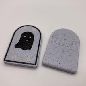 Pode incluir: Duas decora&ccedil;&otilde;es com tema de Halloween. Uma &eacute; uma l&aacute;pide cinza com as letras "R.I.P." e a outra &eacute; uma l&aacute;pide cinza com um gr&aacute;fico de fantasma preto. Ambas t&ecirc;m uma textura salpicada e est&atilde;o sobre um fundo branco.
