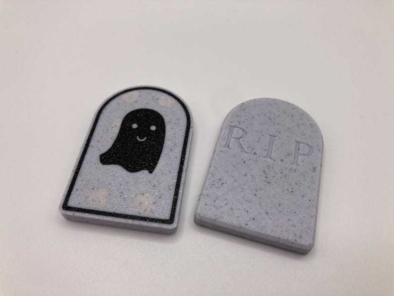 Pode incluir: Duas decora&ccedil;&otilde;es com tema de Halloween. Uma &eacute; uma l&aacute;pide cinza com as letras "R.I.P." e a outra &eacute; uma l&aacute;pide cinza com um gr&aacute;fico de fantasma preto. Ambas t&ecirc;m uma textura salpicada e est&atilde;o sobre um fundo branco.