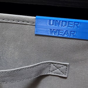 May include: A blue label with the text "UNDERWEAR" on a gray fabric drawer organizer.